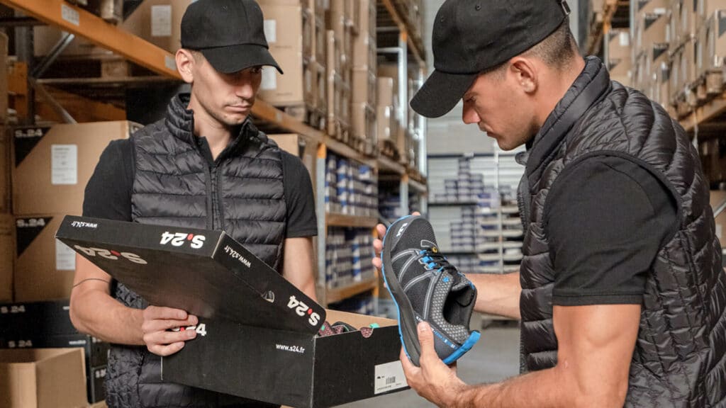deux hommes habillés en noir dans un entrepôt qui tiennent une chaussure et une boite de chaussures dans les mains