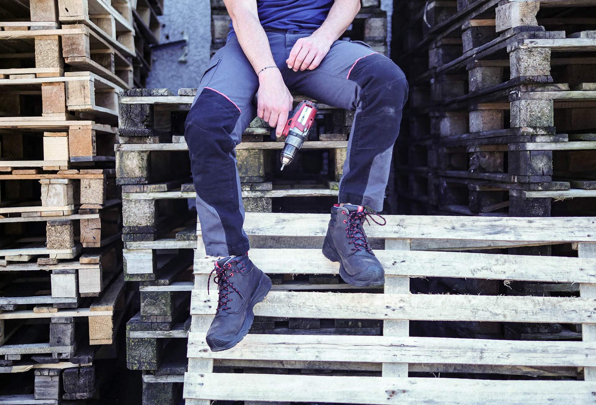 jambes d'un homme assis sur des palettes en bois, tenant dans la main une perceuse