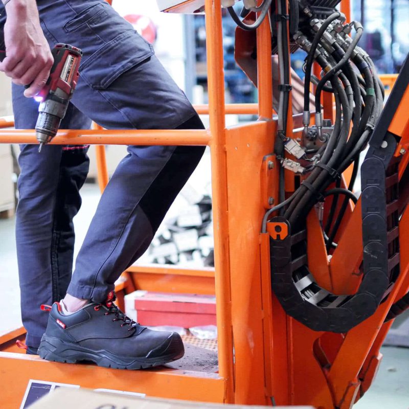 jambes d'un homme sur une nacelle orange tenant dans la main une perceuse