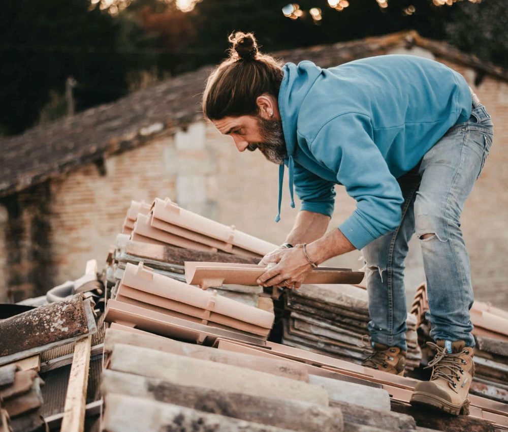 Chaussure de sécurité chantier XPER TP S3 situation homme habillé avec un pull bleu, un jeans et des chaussures marron sur un toit, avec une tuile des les mains