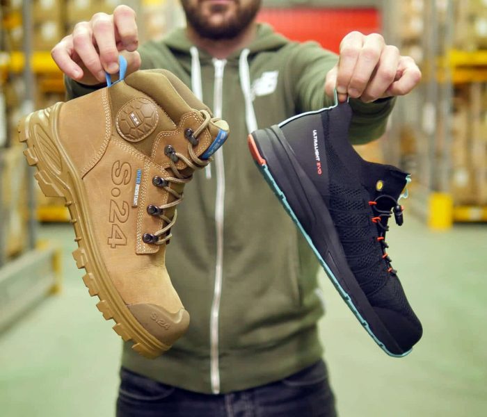 homme tenant au bout des doigts une chaussure de sécurité montante marron et une basket de sécurité noire