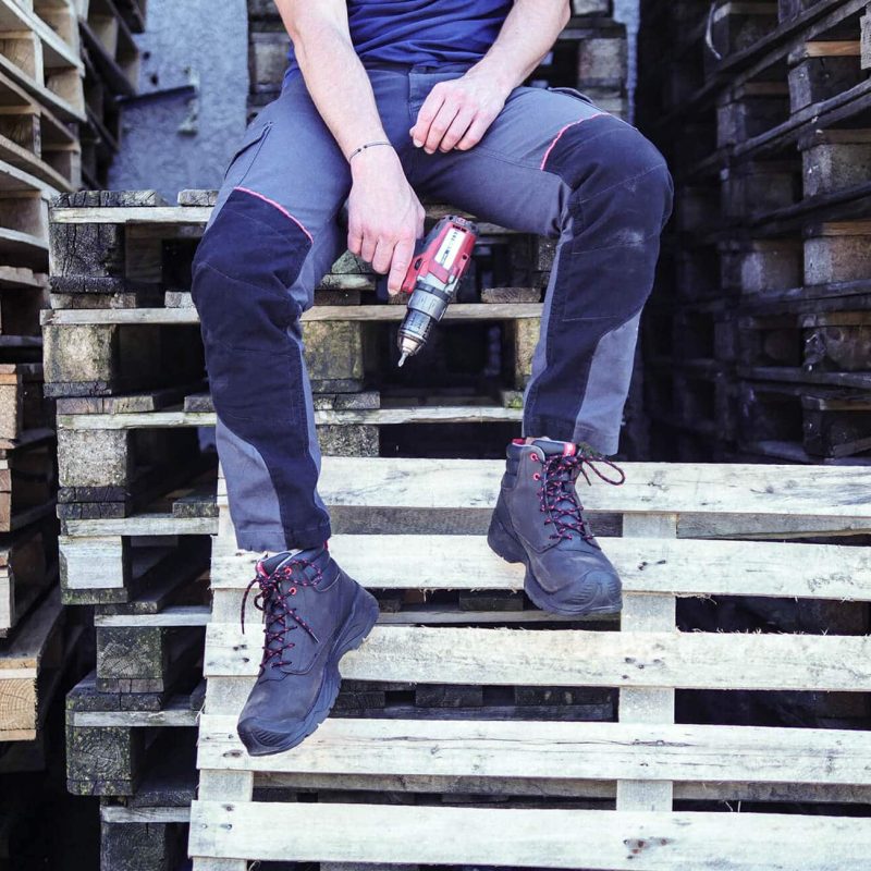 jambes d'un homme assis sur des palettes en bois, tenant dans la main une perceuse