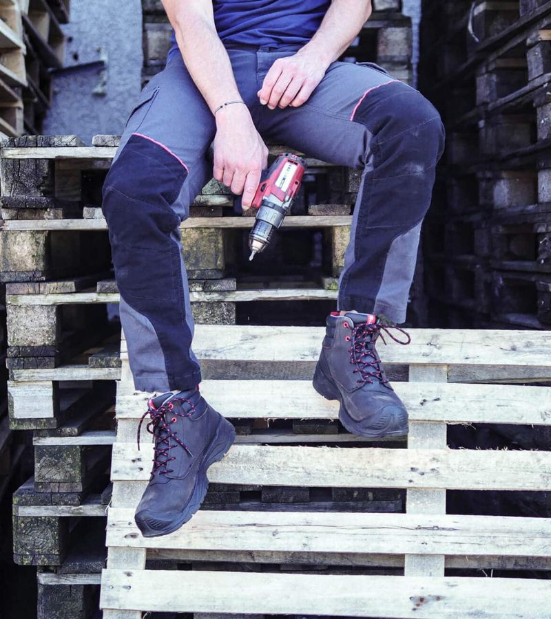 jambes d'un homme assis sur des palettes en bois, tenant dans la main une perceuse