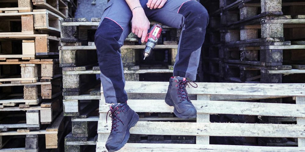 jambes d'un homme assis sur des palettes en bois, tenant dans la main une perceuse