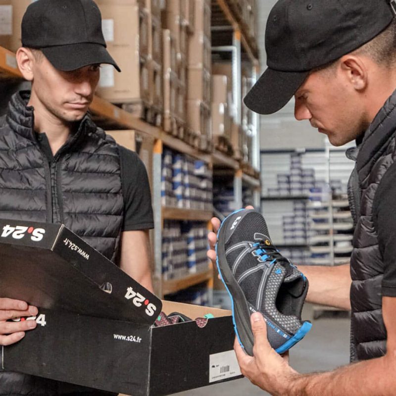 deux hommes habillés en noir dans un entrepôt qui tiennent une chaussure et une boite de chaussures dans les mains