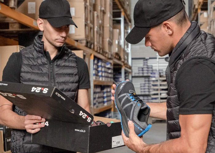 deux hommes habillés en noir dans un entrepôt qui tiennent une chaussure et une boite de chaussures dans les mains