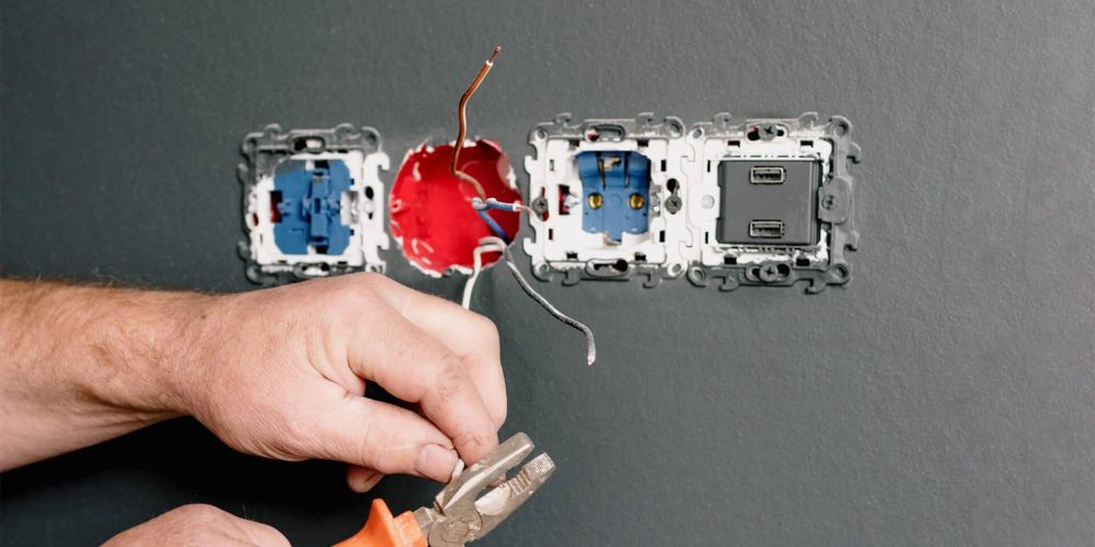 mains d'une personne manipulant les fils électriques d'une prise sur un mur gris