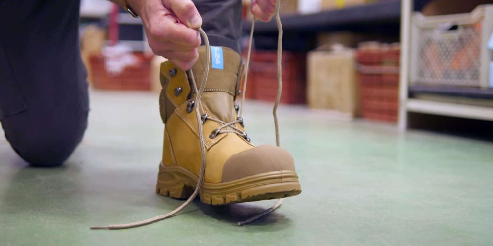 homme à genoux en train de lacer ses chaussures de sécurité marron sur un sol vert