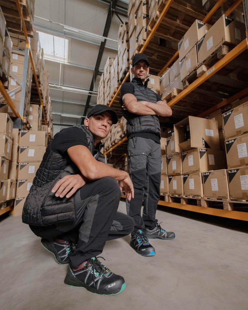 un homme accroupi et un homme debout dans un entrepôt logistique avec des cartons