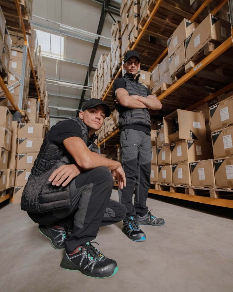 un homme accroupi et un homme debout dans un entrepôt logistique avec des cartons